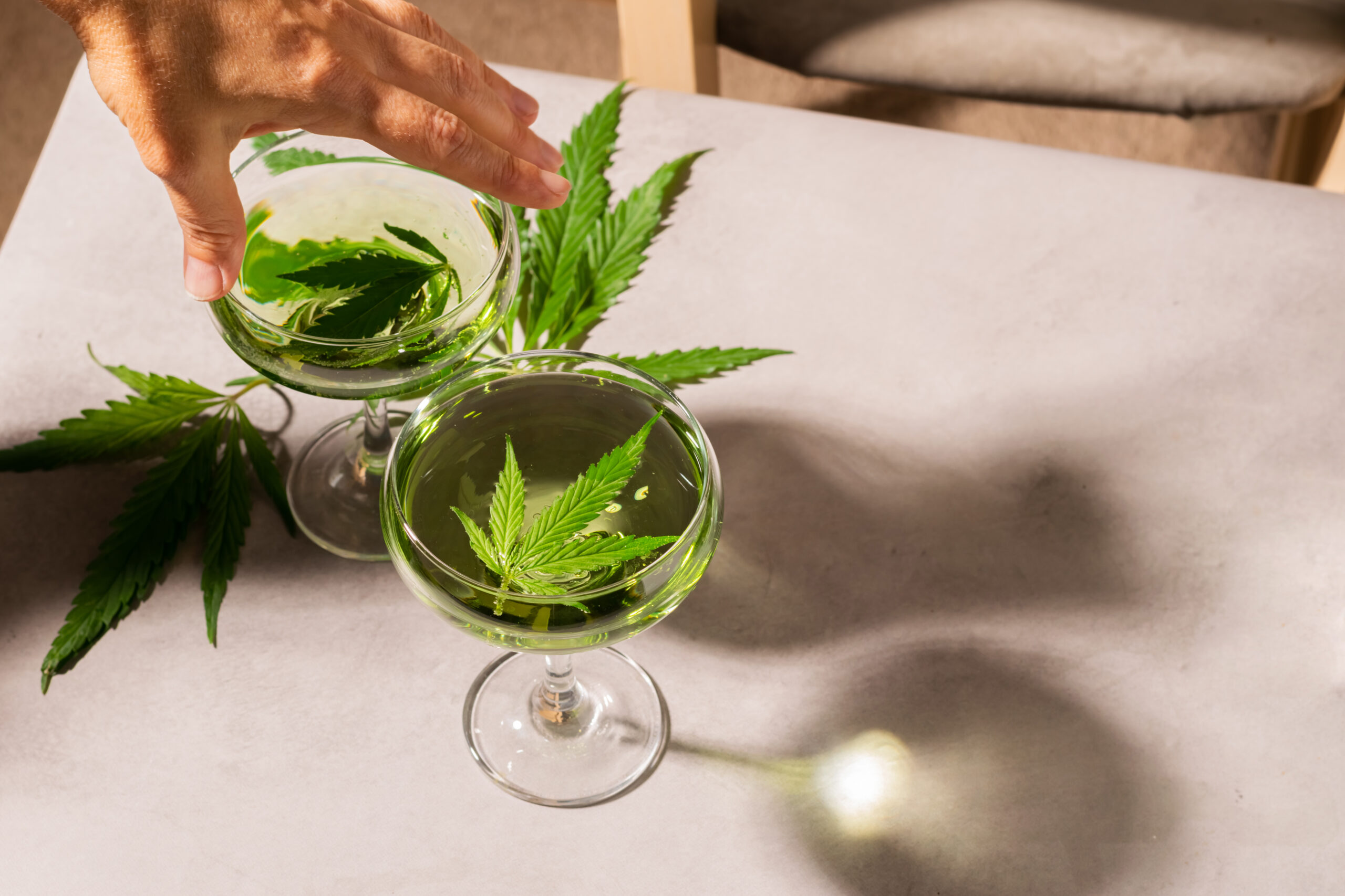 A woman's hand holds glass with green drink from SBD hemp, light concrete background hard shadows